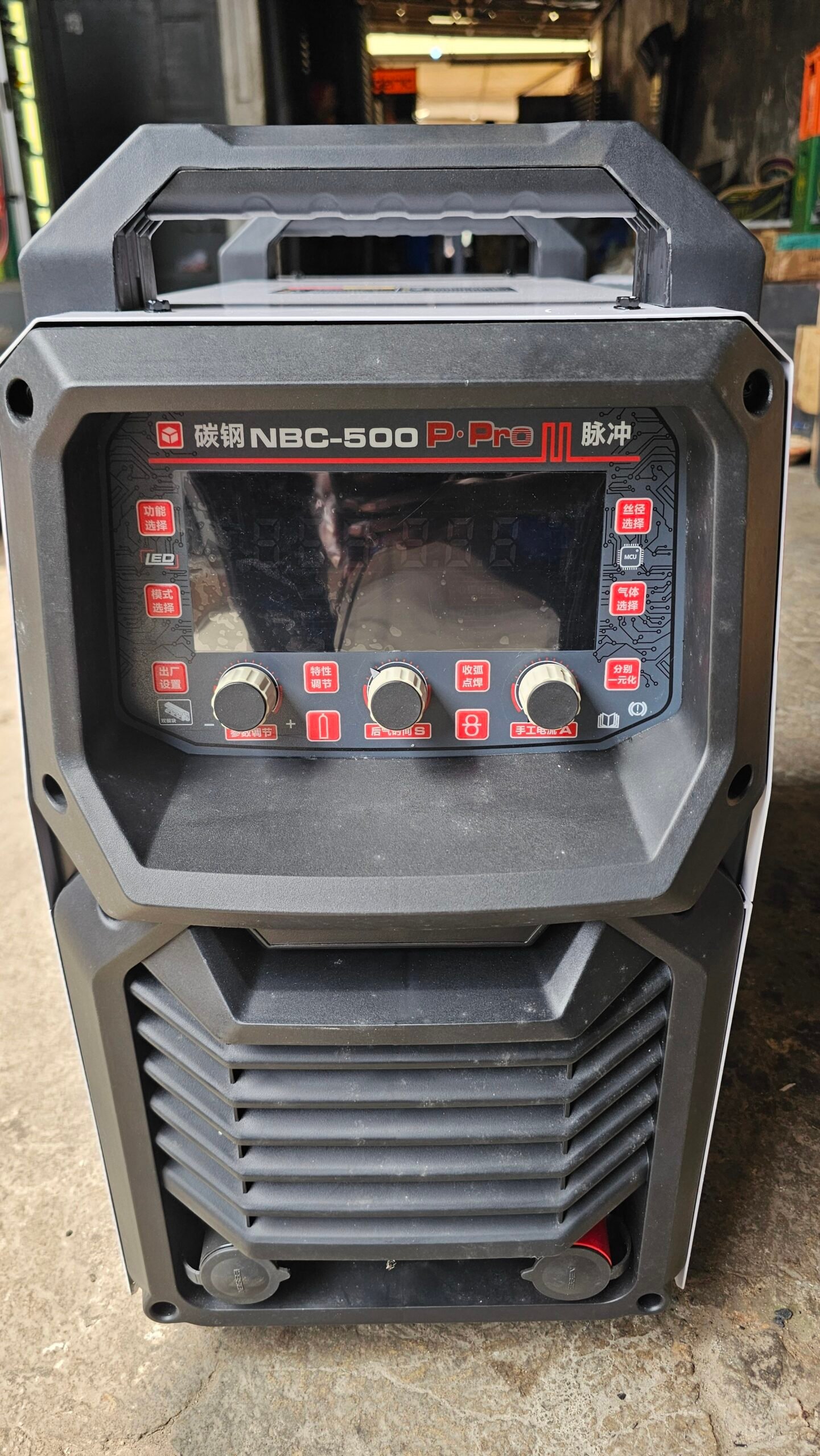 2026/06/1002407399-scaled.jpg - pricemagicgh.com Front view of a NBC-500 P-Pro II welding machine with a digital display, red labeled buttons, and control knobs on a concrete floor.