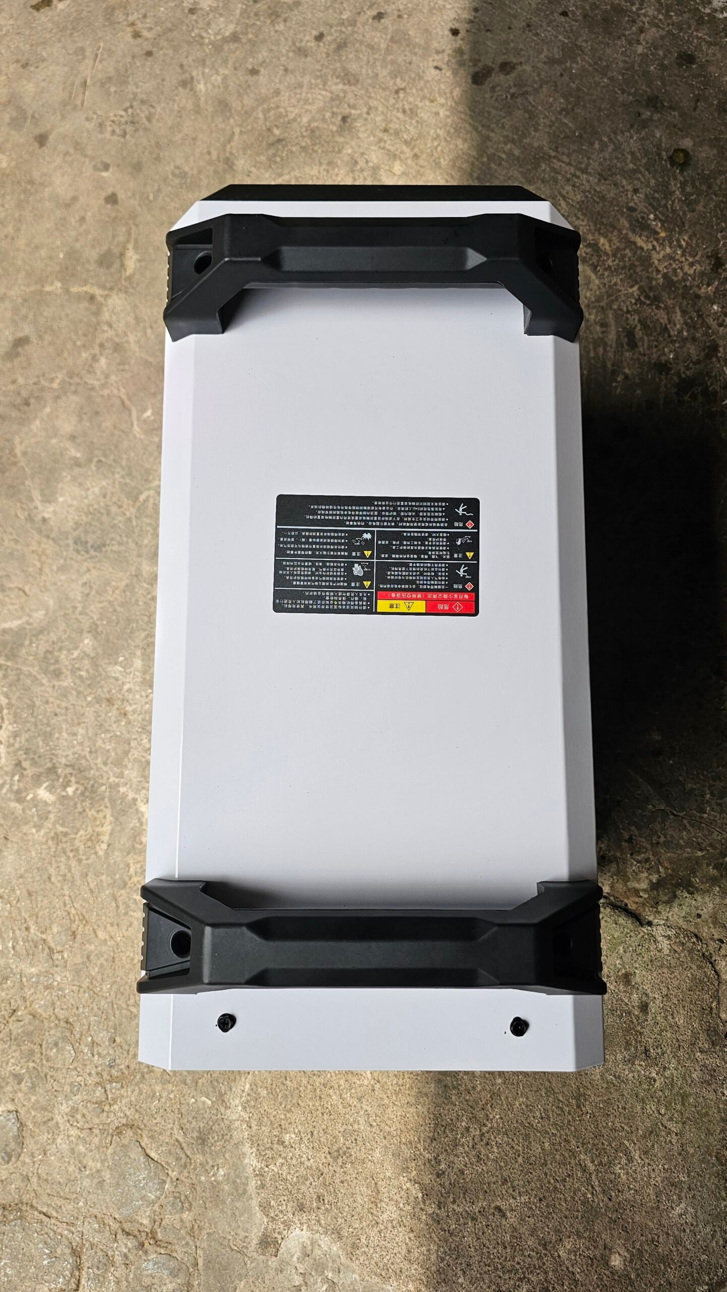 2026/06/1002407403-scaled.jpg - pricemagicgh.com White rectangular battery/module with black protective corner bumpers, lying on a concrete floor; has a warning label on its top surface.