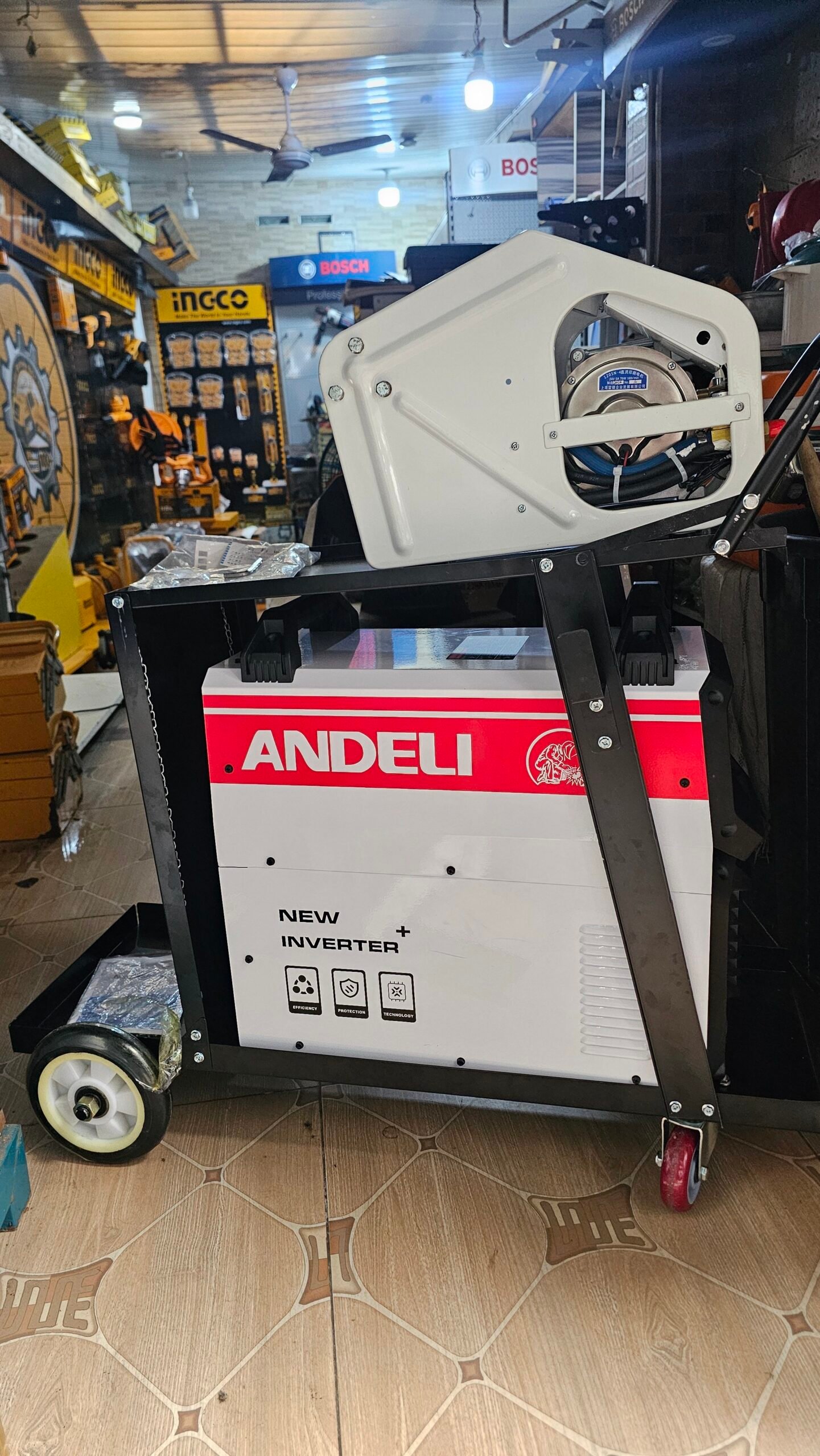 2026/06/1002407952-scaled.jpg - pricemagicgh.com Portable Andeli inverter generator mounted on a wheeled frame in a hardware store, with Bosch and Ingco signs in the background.