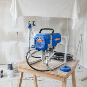 Blue portable paint sprayer on a wooden work table in a dusty painting area under renovation.