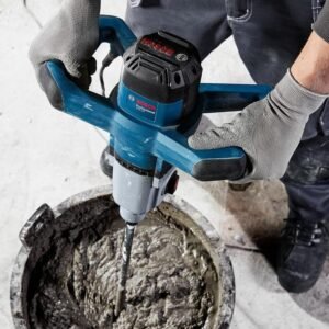 Person wearing gloves operates a blue Bosch concrete drill mixer stirring cement in a bucket surface level view.
