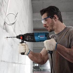 Man wearing safety goggles and gloves drilling into a white concrete wall with a blue corded drill.